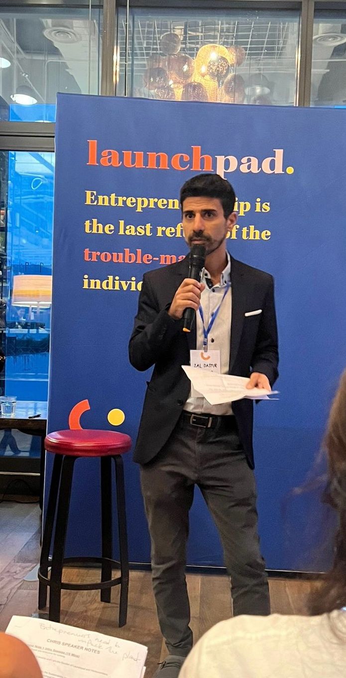 Man in a dark blazer holding a microphone and papers, speaking in front of a blue launchpad banner with motivational text.