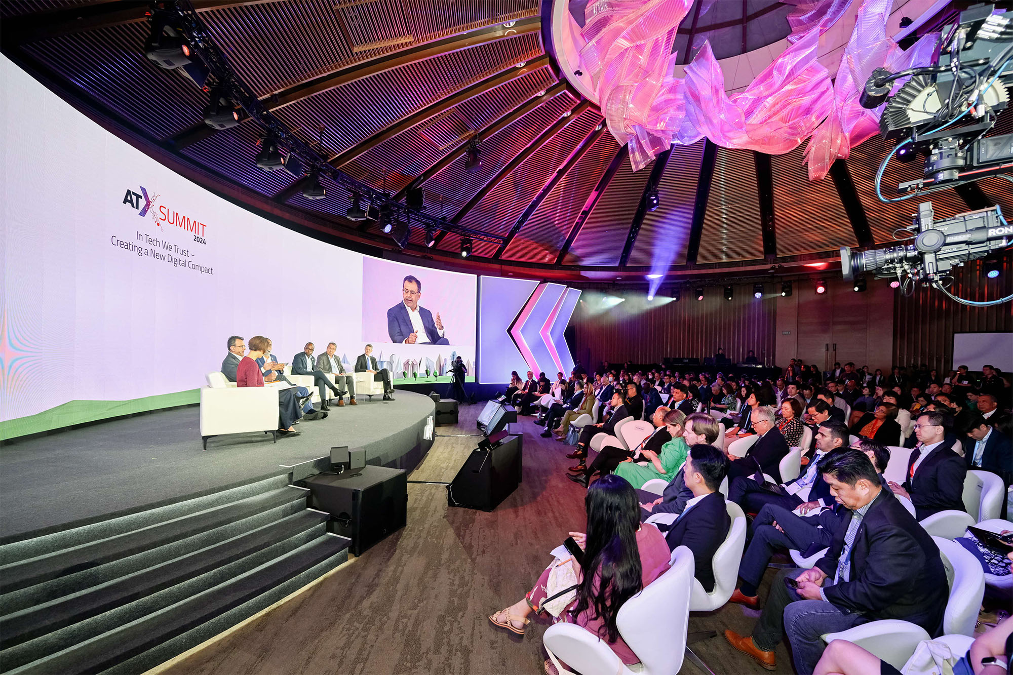 Panel of six speakers seated on a stage at ATX Summit 2024 with a large audience watching in a conference hall.