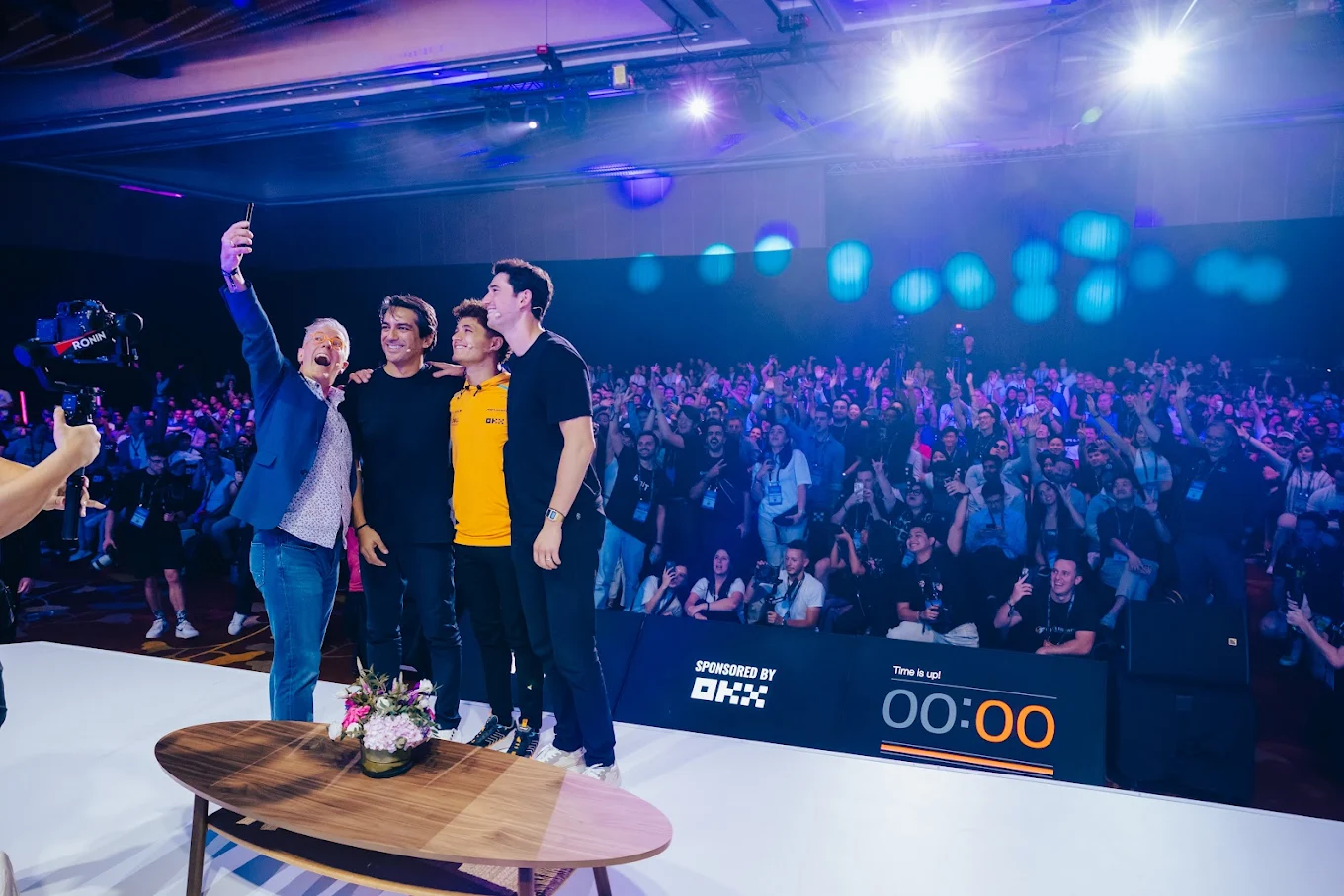 Four men standing on a stage taking a selfie with a large audience visible in the background under bright stage lights.