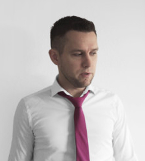 Man with short dark hair wearing a white shirt and a magenta tie looking down and to the side against a plain light background.