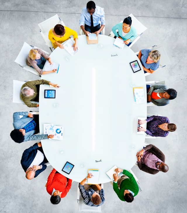 Overhead view of a diverse group of twelve people sitting around an oval white table in a meeting room with documents, tablets, and laptops.