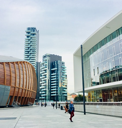 Modern urban plaza with people walking, featuring distinctive tall residential towers and contemporary glass-fronted buildings.