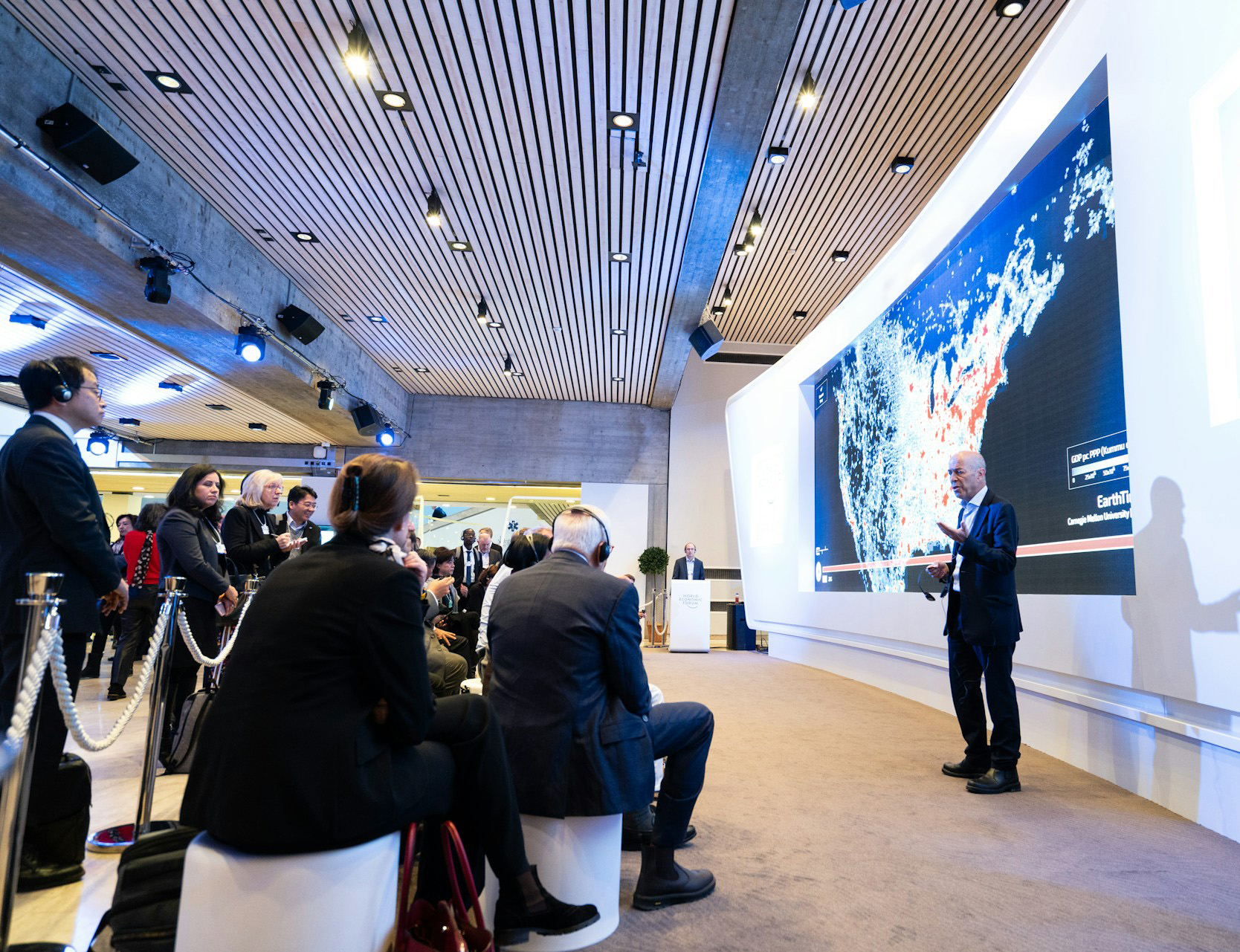 Man in suit giving a presentation in front of a seated audience with a large digital screen displaying a geographic map behind him.