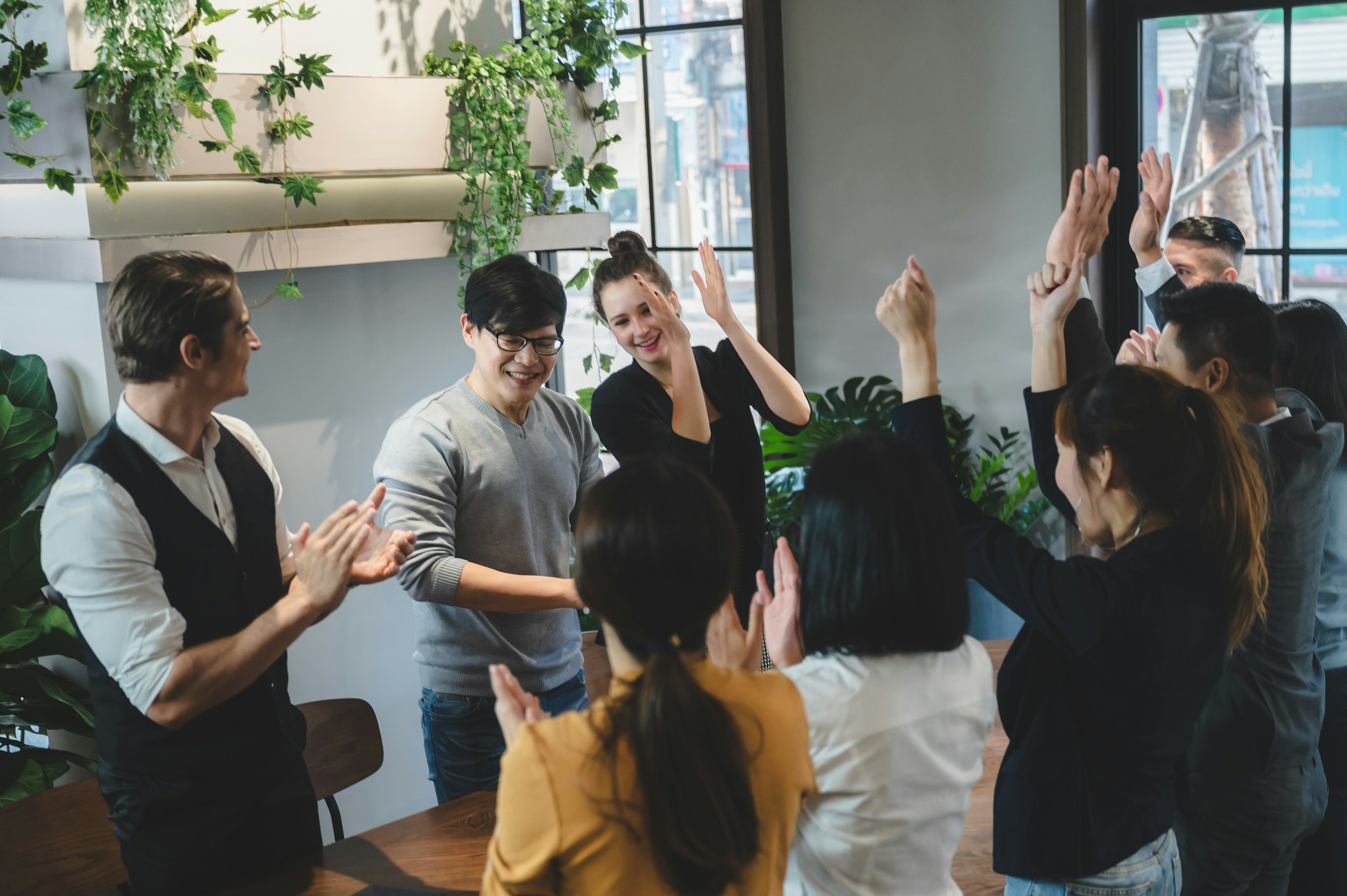 A group of diverse colleagues standing and clapping in an office with plants and large windows.