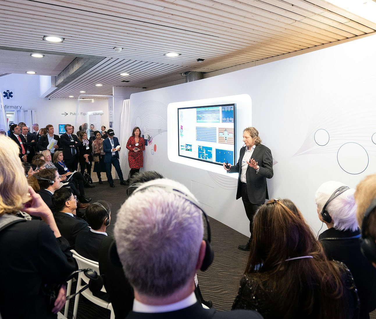 Man in a suit giving a presentation to an audience seated and standing in a modern conference room with a large screen displaying data visuals.