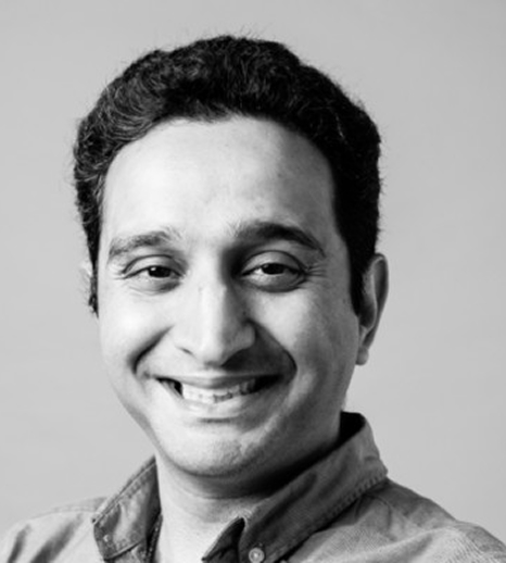 Smiling man with short curly hair wearing a button-up shirt in a black and white portrait.
