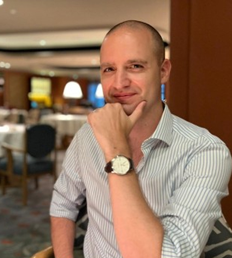 Smiling bald man wearing a striped shirt and watch, sitting indoors with a blurred dining area in the background.