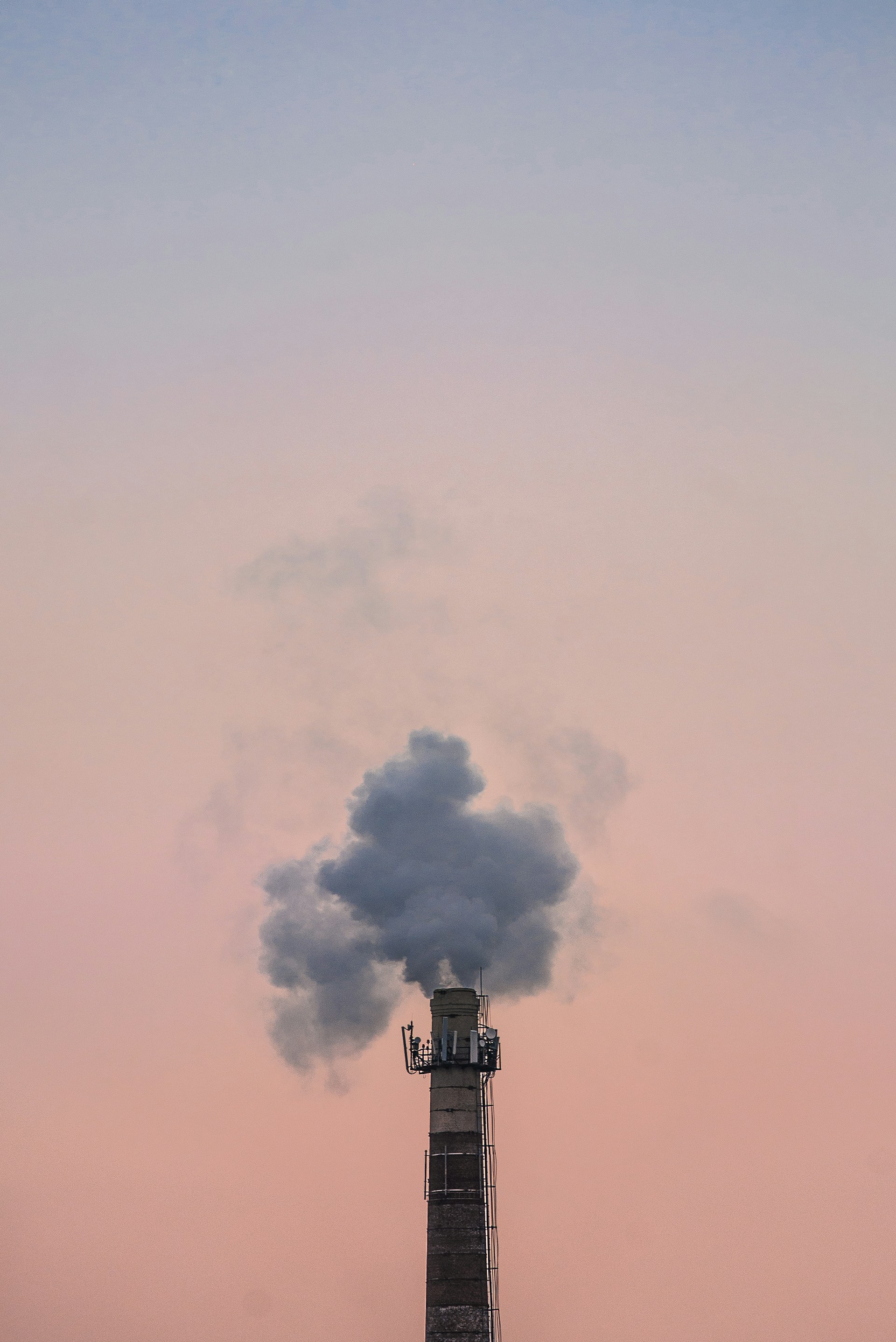 Industrial chimney emitting thick gray smoke against a pastel pink and blue sky.