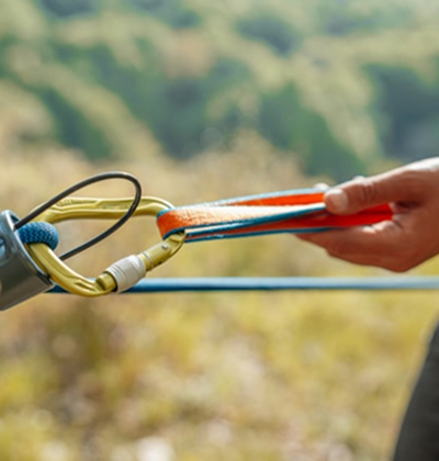 Close-up of a person's hand holding a safety strap connected to a carabiner on a zipline cable outdoors.