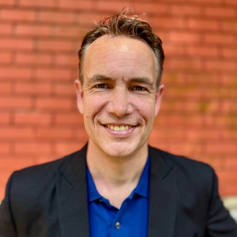 Smiling middle-aged man with short brown hair wearing a blue shirt and dark blazer, standing in front of a red brick wall.