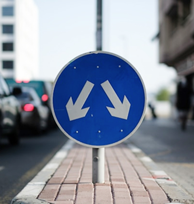 Blue traffic sign with two white arrows pointing diagonally downward to the left and right on a median strip between traffic lanes.