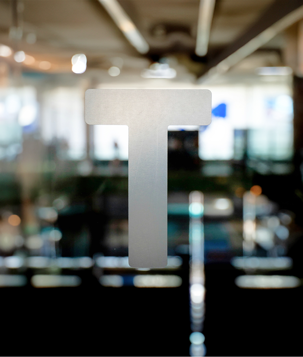 Large frosted letter T on a glass door with a blurred office background.