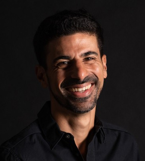 Smiling man with short curly hair and a beard wearing a black shirt against a dark background.